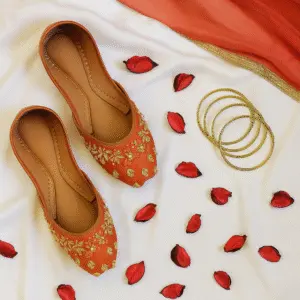Orange embroidered khussa shoes for women with gold detailing, traditional handmade ethnic footwear styled with rose petals and gold bangles for bridal and festive wear.