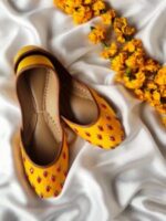 Mustard yellow embroidered khussa jutti placed on white fabric with yellow flowers, Pakistan product photo