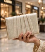 Hand holding ivory party wear clutch bag with gold clasp and bangles