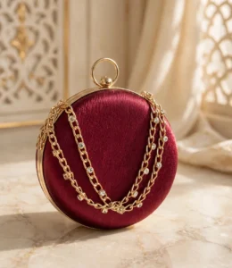 Maroon round clutch bag with gold ring handle and rhinestone chain on marble